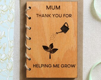 May include: Wooden card with a brown background and a light brown wooden front. The front has a black and white illustration of a watering can pouring water onto a plant. The text on the card reads "Mum Thank You For Helping Me Grow".