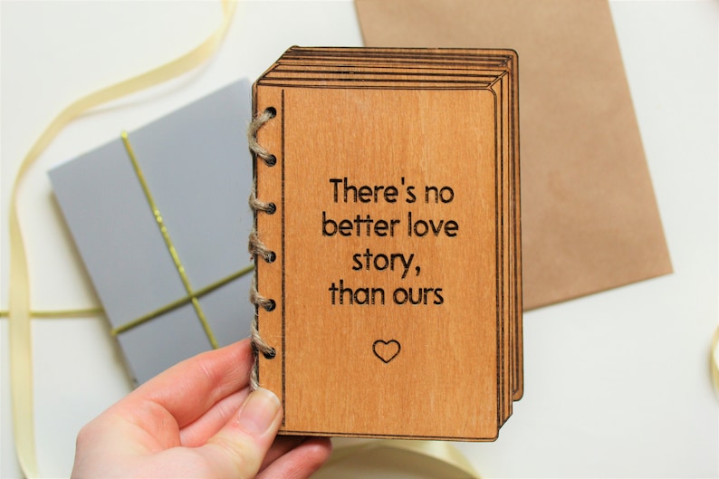 Puede incluir: Un libro de madera con un mensaje que dice "There's no better love story, than ours" con un peque&ntilde;o s&iacute;mbolo de coraz&oacute;n en la parte inferior. El libro est&aacute; encuadernado con cuerda.