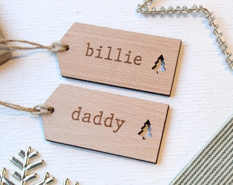 Könnte beinhalten: Zwei Holz-Geschenkanhänger mit den eingravierten Namen "billie" und "daddy". Jeder Anhänger hat einen kleinen Ausschnitt in Form eines Weihnachtsbaums. Die Anhänger sind mit Bindfaden befestigt.