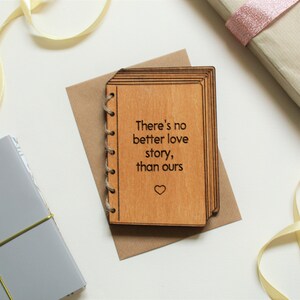Puede incluir: Una tarjeta de madera con forma de libro con el texto "There's no better love story, than ours" y un peque&ntilde;o s&iacute;mbolo de coraz&oacute;n grabado en la parte delantera.