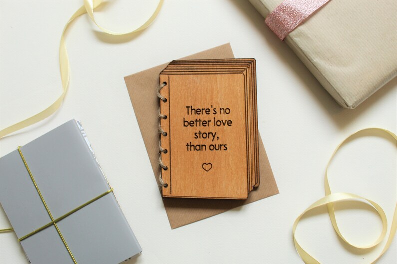 Puede incluir: Una tarjeta de madera con forma de libro con el texto "There's no better love story, than ours" y un peque&ntilde;o s&iacute;mbolo de coraz&oacute;n grabado en la parte delantera.