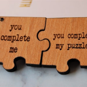 以下が含まれることがあります： 「you complete me」と「you complete my puzzle」と書かれた木製のジグソーパズル2ピース。ピースは銀色のチェーンでつながっています。