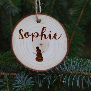 Puede incluir: Un adorno redondo de madera con el nombre "Sophie" grabado en él. También se graba una silueta de muñeco de nieve en el adorno.