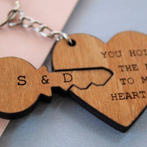 Pode incluir: Chaveiros de madeira com design de chave e cora&ccedil;&atilde;o. A chave tem as iniciais "S & D" gravadas, e o cora&ccedil;&atilde;o diz "YOU HOLD THE KEY TO MY HEART". Os chaveiros s&atilde;o conectados por uma corrente prateada.