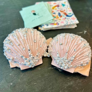 Könnte beinhalten: Ein Paar hellrosa muschelförmige Pasties, verziert mit weißen und blauen Perlen und Perlen. Die Pasties befinden sich auf einem dunklen Hintergrund, mit Servietten im Hintergrund.