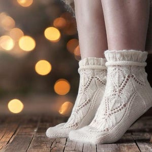 May include: A pair of off-white, knitted socks with a decorative openwork pattern. The socks have a ruffled cuff and are worn on a wooden floor. The background features soft, blurred golden lights.