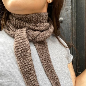 May include: A brown knitted scarf is wrapped around the neck. The scarf is made of a textured yarn and has long, dangling ends. The scarf is worn over a gray t-shirt. The background is a dark, textured surface.