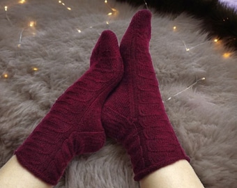 May include: Burgundy-coloured knitted socks with a textured wave pattern. The socks are made of a soft material. The socks are shown on a fluffy, light-coloured rug. These socks are designed to keep feet warm.