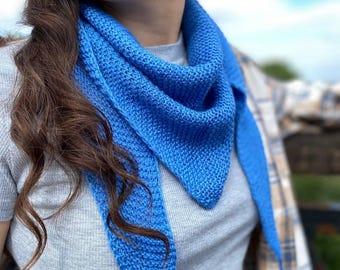Könnte beinhalten: Ein leuchtend blauer Strickschal in Dreiecksform. Der Schal besteht aus einem strukturierten Strickstoff und wird um den Hals gelegt. Die leuchtend blaue Farbe kontrastiert mit dem grauen Oberteil und dem Himmel im Hintergrund.