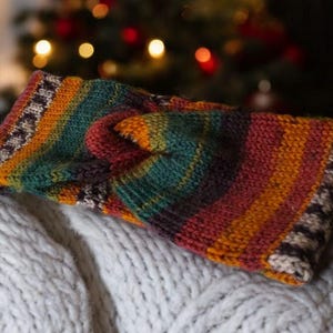 May include: A knitted headband in a variety of colours, including orange, green, red, and brown stripes. The ends of the headband have a patterned border. The headband is resting on a white, textured surface.