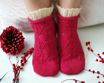 May include: A pair of bright pink knitted socks with a diamond pattern. The socks have a cream-colored, ruffled cuff. Red berries and a decorative red ball are in the background.