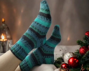 May include: A pair of teal and black striped socks with a fuzzy texture. The socks are worn on a person's feet and are resting on a white surface. The background includes a lit candle and a Christmas tree with red ornaments.