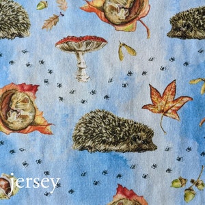 May include: A blue fabric with a repeating pattern of hedgehogs, mushrooms, leaves, and acorns. The fabric has the word "jersey" printed on it.