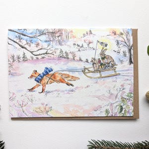 Könnte beinhalten: Aquarellillustration eines Fuchses, der einen Schlitten mit einem Kaninchen und anderen Tieren durch eine verschneite Landschaft zieht. Der Fuchs trägt einen blauen Schal. Der Schlitten hat eine Flagge. Die Karte hat einen braunen Rand.