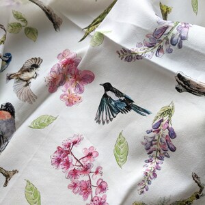 May include: White fabric with a repeating pattern of pink flowers, green leaves, and birds. The birds are black and white, blue and white, and green and yellow. The text "poplin" is printed in black at the bottom of the fabric.