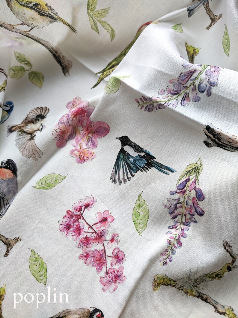 May include: White fabric with a repeating pattern of pink flowers, green leaves, and birds. The birds are black and white, blue and white, and green and yellow. The text "poplin" is printed in black at the bottom of the fabric.