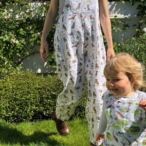 May include: Two people wearing white overalls with a colorful print of dogs and other animals. The overalls have adjustable straps and pockets. The background is a green grassy area.