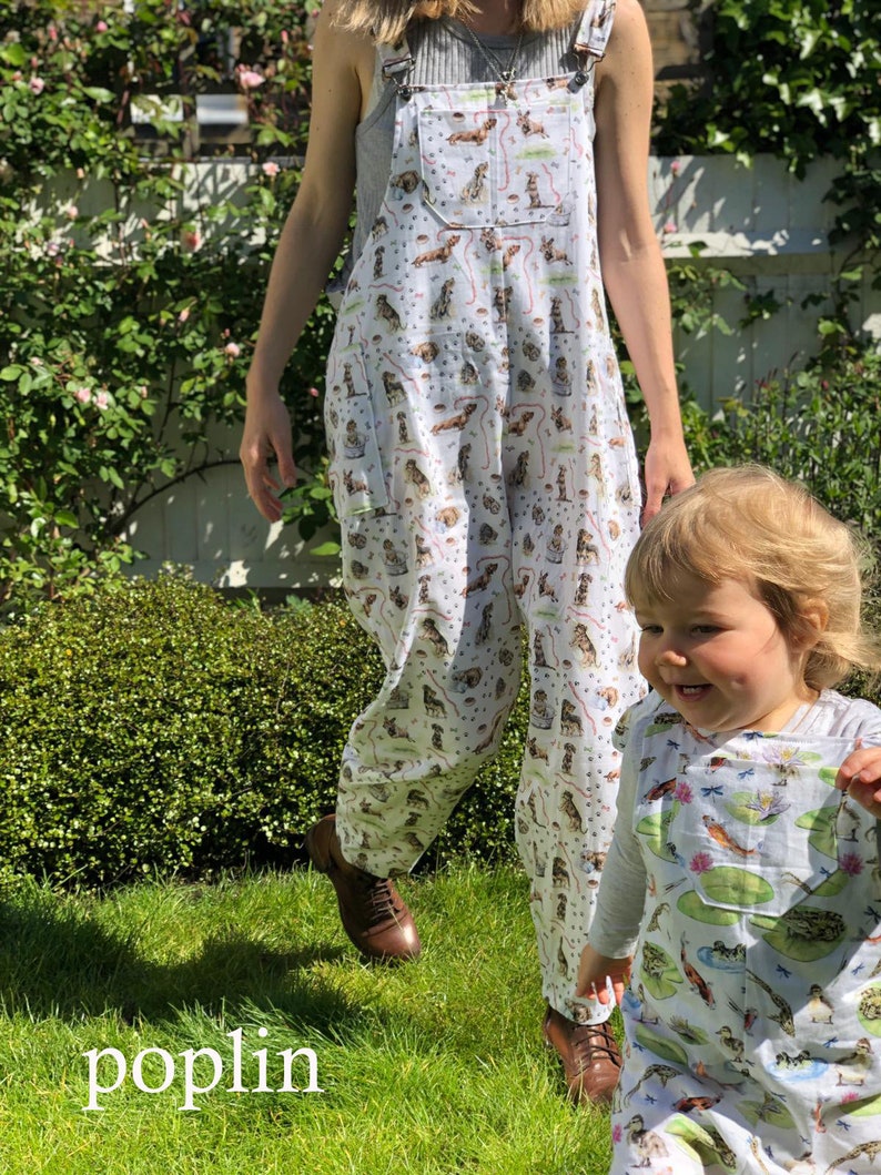 May include: Two people wearing white overalls with a colorful print of dogs and other animals. The overalls have adjustable straps and pockets. The background is a green grassy area.
