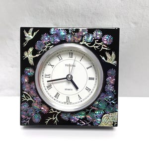 May include: Square black quartz clock with a white face and Roman numerals. The clock is decorated with iridescent floral and bird designs. The clock face reads "Yusung Quartz".