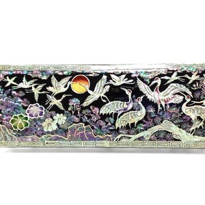 May include: A rectangular black lacquer box featuring an intricate mother-of-pearl inlay design. The scene depicts multiple cranes in flight and standing among stylised flowers, rocks, and a circular sun, all framed by a geometric border pattern.