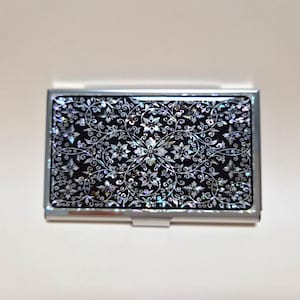 Könnte beinhalten: Ein rechteckiges Visitenkartenetui aus Metall mit einem polierten silbernen Rahmen und einer schwarzen Einlage, die mit einem komplizierten, irisierenden Blumen- und Rankenmuster aus Perlmutt verziert ist, das die gesamte Vorderseite bedeckt.