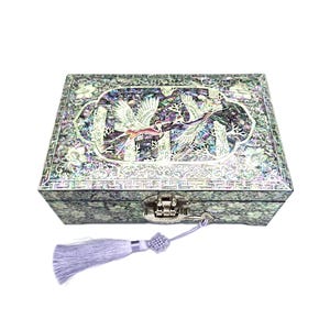 May include: A rectangular jewelry box with a mother-of-pearl inlay. The box features a decorative design with birds and floral patterns. It has a silver-colored clasp and a matching tassel. The box is closed and sits on a white background.