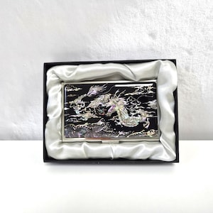 Może przedstawiać: Czarny wizytownik z motywem smoka. Smok jest misternie zdobiony iryzującymi akcentami, na czarnym tle. Wizytownik jest prezentowany w czarnym pudełku z satynową wyściółką.