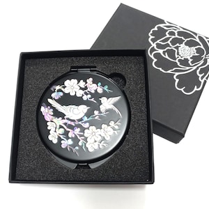May include: Black compact mirror with a bird and floral design. The mirror is round and features two birds perched on a flowering branch. The mirror is in a black box with a floral design on the lid.
