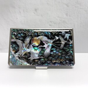 May include: A rectangular, silver-toned business card holder with an iridescent, black, and green inlaid design. The design features flying cranes, a full moon, and stylized trees. The card holder is set against a white background.