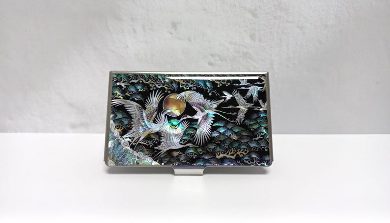 May include: A rectangular, silver-toned business card holder with an iridescent, black, and green inlaid design. The design features flying cranes, a full moon, and stylized trees. The card holder is set against a white background.