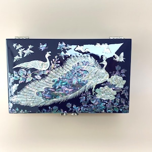 Puede incluir: Una caja de joyería rectangular azul marino con un diseño decorativo de pavo real y flores. Las plumas de la cola del pavo real son iridiscentes, con detalles en blanco y azul. También se presentan mariposas y flores.