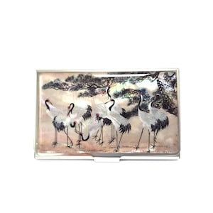 May include: A rectangular business card holder with a mother-of-pearl finish. The holder features a painting of several cranes with black and white feathers, standing in a field with a tree in the background.