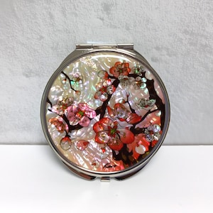 Op de afbeelding: Een ronde, zilveren compact spiegel met een bloemenmotief. Het ontwerp toont een tak met roze en rode bloemen met groene bladeren op een witte en iriserende achtergrond.