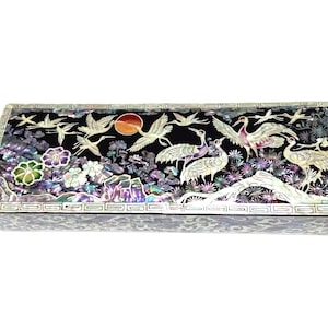 May include: Rectangular black box with a mother-of-pearl inlay design. The design features cranes, flowers, and a setting sun. The box has a decorative border and is set against a white background.