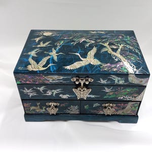 Pode incluir: Uma caixa de joias retangular azul-escura com incrustações de madrepérola iridescente de grous, árvores e nuvens. A caixa inclui uma tampa articulada com um fecho de metal em forma de borboleta e duas gavetas inferiores com puxadores.