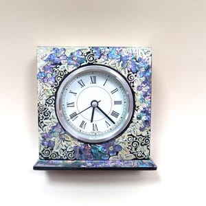 May include: A square decorative clock with a white clock face and black Roman numerals. The clock is framed with a floral design in shades of blue, purple, and white. The clock has a small shelf at the bottom.