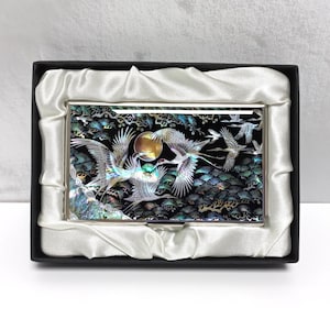 May include: A silver business card holder with a mother-of-pearl inlay featuring a crane design. The cranes are depicted in flight against a dark background with a full moon. The holder is presented in a black box with a white satin lining.