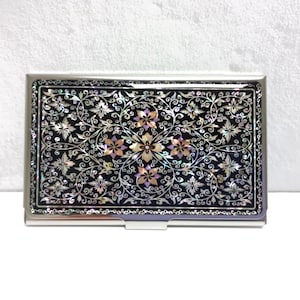 Puede incluir: Un porta tarjetas de plata con fondo negro y un diseño floral hecho de nácar iridiscente.