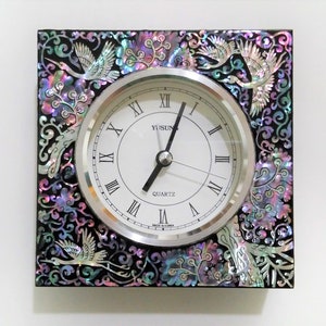 May include: A square clock with a black frame and a white face. The frame is decorated with iridescent mother-of-pearl inlay in a floral pattern. The clock face has Roman numerals and the words "YUSUN QUARTZ" printed on it.