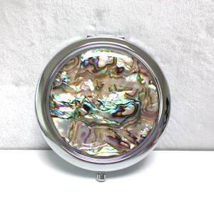 May include: A circular, silver compact mirror with an abalone shell design. The shell has iridescent colours of green, blue, and gold. The compact is closed and rests on a white surface.