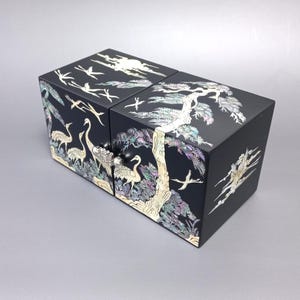 May include: A black rectangular jewelry box featuring intricate mother-of-pearl inlay designs of cranes and pine trees on the top and front panels. The box includes two front-facing drawers with small round knobs and a smooth, dark finish.