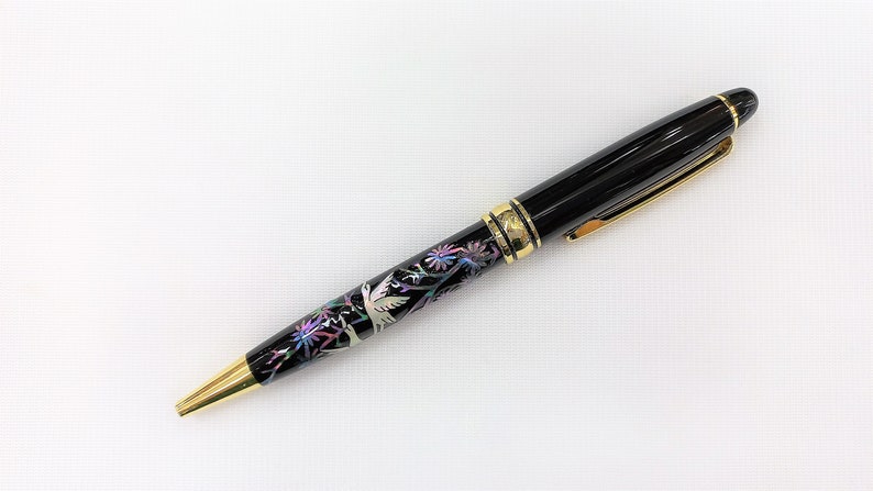 May include: Black ballpoint pen with gold accents. The pen features a decorative band with a floral and bird design in iridescent colors. The pen's tip and clip are gold-colored.