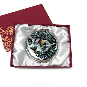 May include: A round, silver compact mirror with a nature scene of white birds and a golden sun, presented in a red gift box with a white satin lining. The box is open, revealing the mirror.