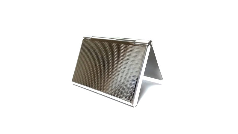 May include: A silver-colored business card holder with a textured surface. The holder is in a triangular shape, with a hinged top and a reflective finish. The design is simple and modern, suitable for professional use.