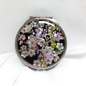 Könnte beinhalten: Ein silberner Kompakt-Spiegel mit schwarzem Hintergrund und einem Design aus rosa und weißen Blumen aus Perlmutt.