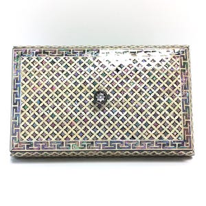 Pode incluir: Caixa retangular com incrustações decorativas de madrepérola. A caixa apresenta um padrão de treliça geométrica na tampa e uma borda decorativa. Um pequeno botão ornamentado serve como puxador. A caixa está sobre um fundo branco.