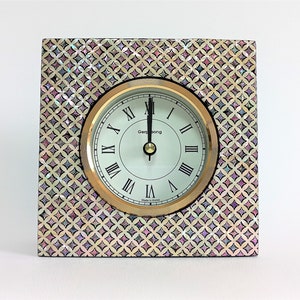 May include: A square clock with a white face and black Roman numeral hour markers. The clock has a gold-coloured frame and is set against a background of iridescent mother-of-pearl tiles. The clock face reads "Gemstone" and "Made in Korea".