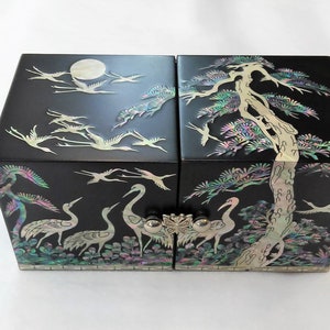 May include: Black lacquered box with iridescent mother-of-pearl inlay. The box features a design of cranes, a full moon, and a tree. The box has a hinged opening and decorative metal hardware. The box is likely used for jewelry or keepsakes.