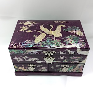 May include: A rectangular purple jewelry box featuring iridescent mother-of-pearl inlay depicting cranes, pine trees, and clouds. The box includes a hinged lid, two lower drawers, and a silver-toned butterfly-shaped metal latch on the front.