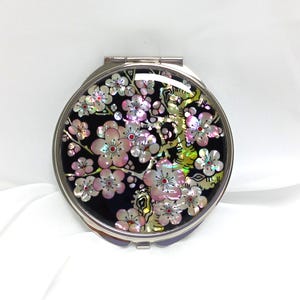 Peut inclure: Un miroir de poche rond et argenté avec un fond noir. Le miroir est orné d'un motif floral avec des fleurs roses et blanches. Les fleurs ont des reflets irisés, et le boîtier a une ouverture à charnière.